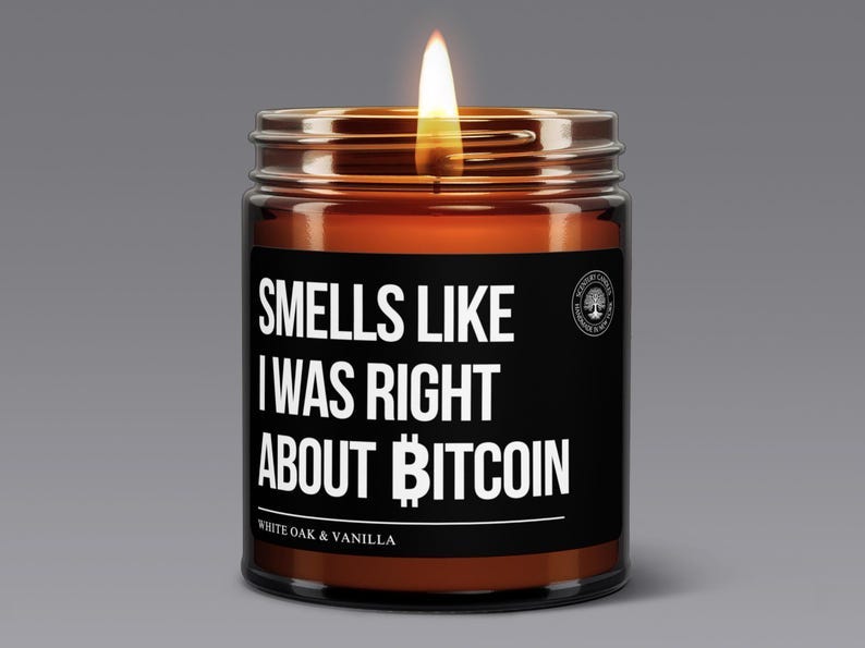 May include: A brown amber glass candle with a black label. The label features white text that reads: 'SMELLS LIKE I WAS RIGHT ABOUT BITCOIN.' Below this, in a smaller font, it says 'WHITE OAK & VANILLA'. The candle is lit, with a small flame visible at the top. The overall theme is humorous and related to cryptocurrency.