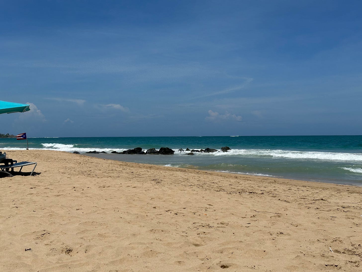 Picture of a beach with waves, a few rocks, and a bright blue sky 