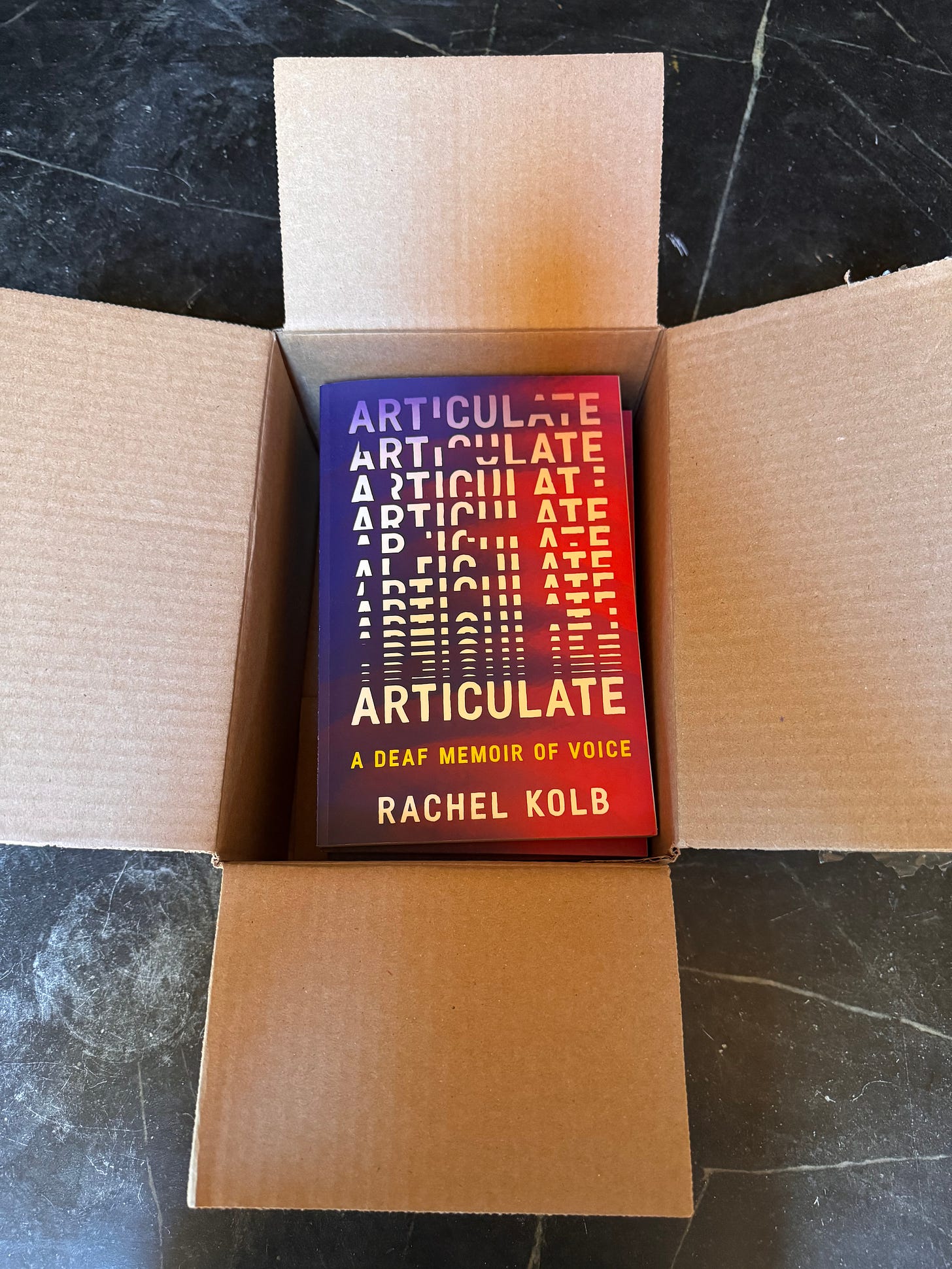 An open cardboard box on a counter, stacked copies of ARTICULATE: A DEAF MEMOIR OF VOICE inside. White letters assemble across a swirling pink-and-purple background.