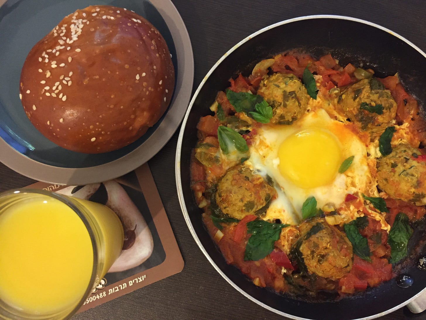 A pan of shakshuka with a sunny-side-up egg in the center, surrounded by fish balls in a rich tomato sauce with fresh basil. Served alongside a round challah bun and a glass of orange juice. Part of Liat Portal’s Foodie Disorder journey on The Liat Show.