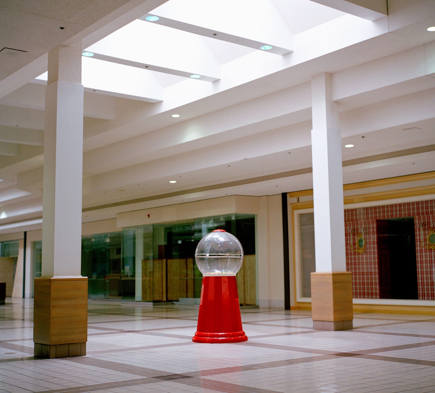 A large empty gumball machine sits in the corridor of a deserted mall.