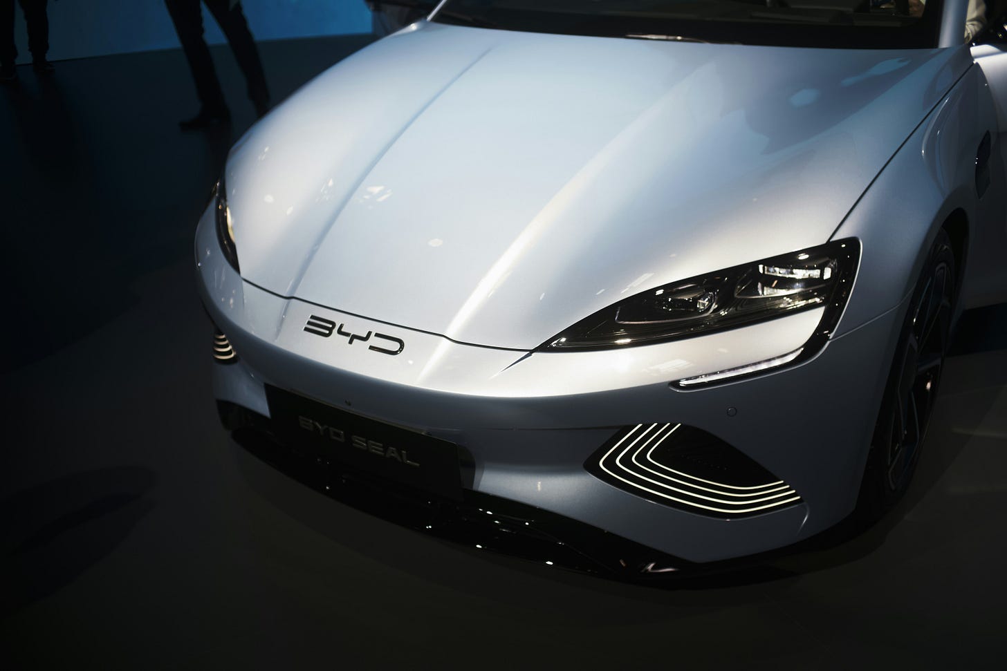 foto mostrando o capô de um carro da BYD com o logo da fabricante. carro prateado com fundo escuro e parachoque preto foto mostrando o capô de um carro da BYD com o logo da fabricante. carro prateado com fundo escuro e parachoque preto