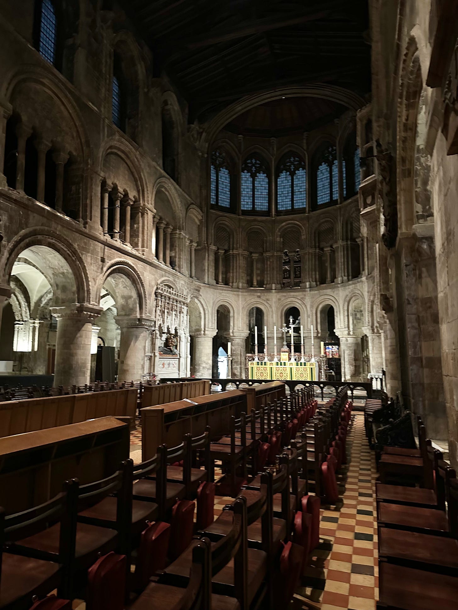Interior of a large historic cathedral with Romanesque arches, stone columns, rows of wooden chairs, and a softly lit altar beneath tall stained-glass windows.
