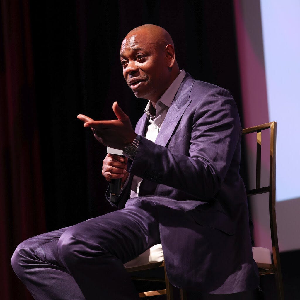 Dave Chappelle sits on a chair on a stage holding a microphone.