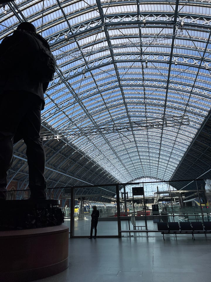Outside image of the station, inside the station on the first floor, high vaulted glass roof over station end of line stops, a close up of a bronze statue with a half a face and a 3D tunnel and arches, photo of the large bronze sculpture of a man and women hugging