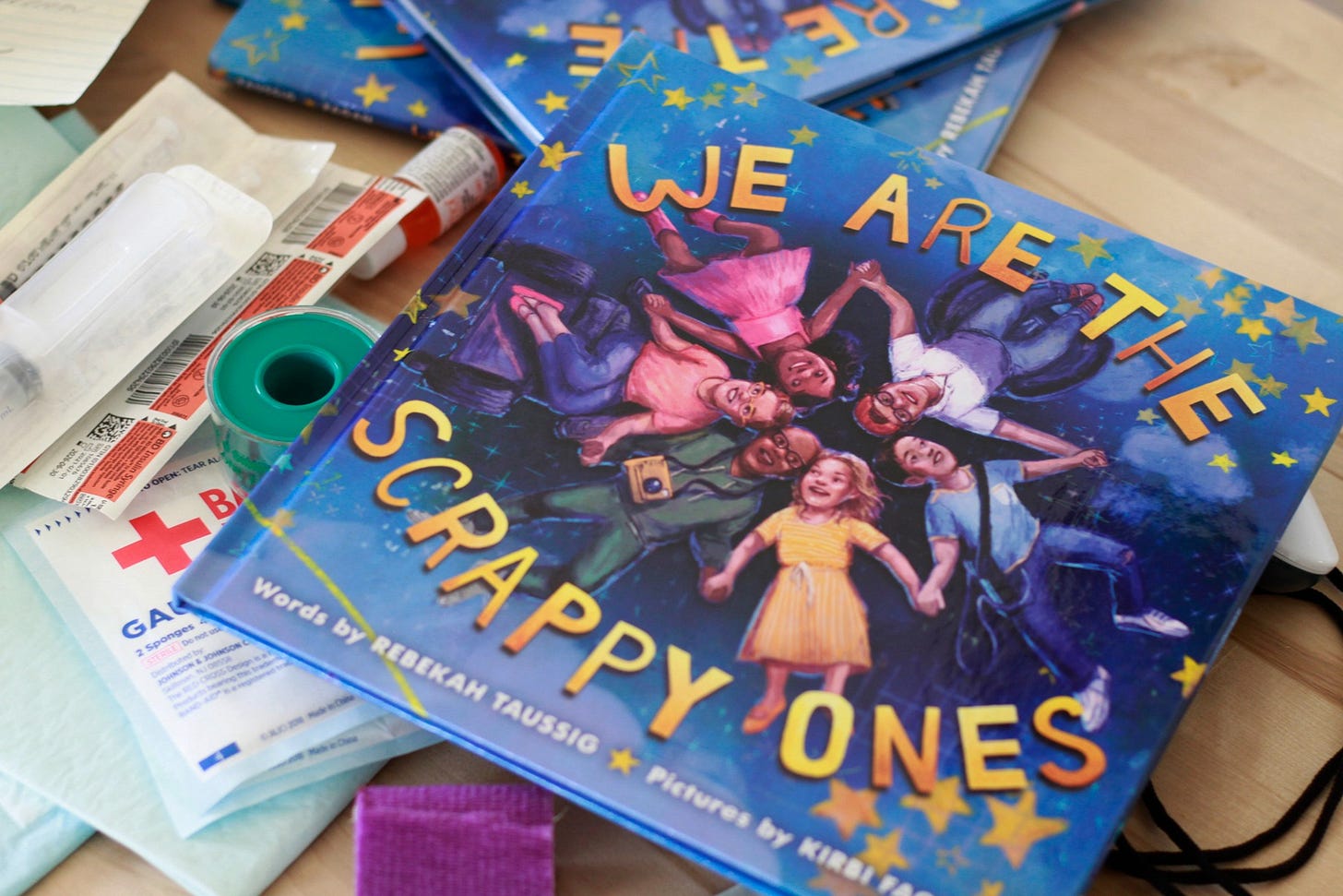 The book We Are the Scrappy Ones sits on a table covered in medical supplies. On the cover, a group of kids hold hands making the shape of a star between them. They almost look like they're floating in outer space surrounded by yellow stars. The book We Are the Scrappy Ones sits on a table covered in medical supplies. On the cover, a group of kids hold hands making the shape of a star between them. They almost look like they're floating in outer space surrounded by yellow stars.
