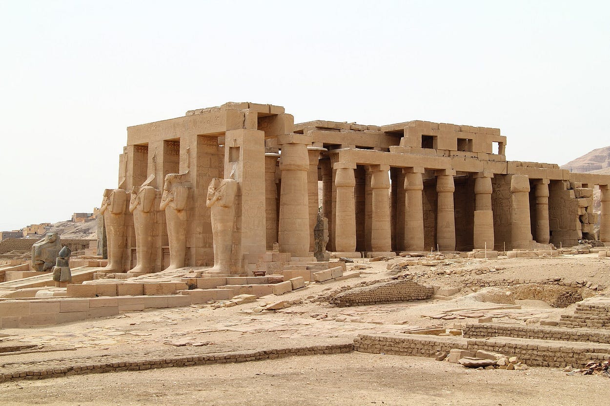 A medium-distance view of an ancient pillared temple in a desert setting with four large statues guarding the entrance. A medium-distance view of an ancient pillared temple in a desert setting with four large statues guarding the entrance.