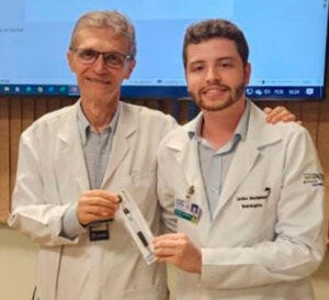 Dois homens usando jalecos brancos estão lado a lado em um ambiente hospitalar ou acadêmico. Uma delas entrega um pequeno objeto à outra, enquanto ambas sorriem para a câmera Dois homens usando jalecos brancos estão lado a lado em um ambiente hospitalar ou acadêmico. Uma delas entrega um pequeno objeto à outra, enquanto ambas sorriem para a câmera