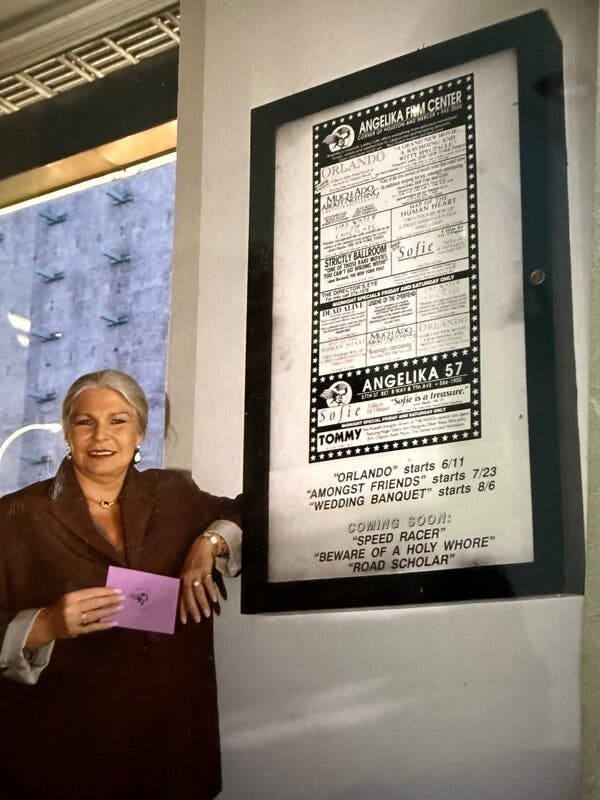 A blonde woman in a brown blazer smiles, posing next to a poster listing movie showtimes.
