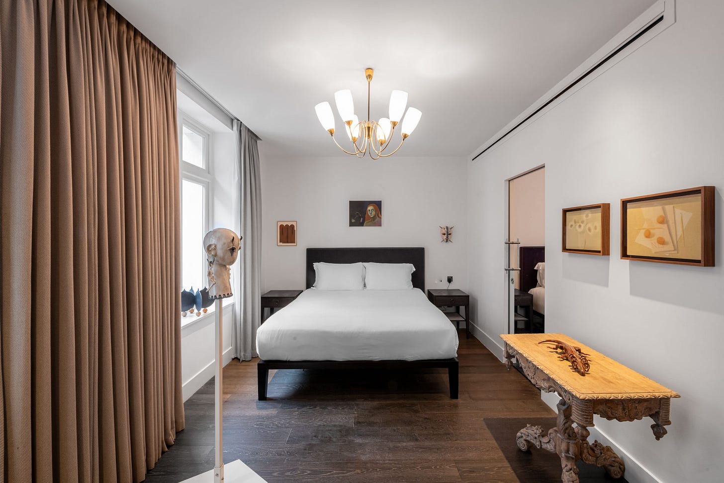 Bright hotel bedroom installation with a made bed, chandelier, heavy curtain, and framed artworks staged like a lived-in room.