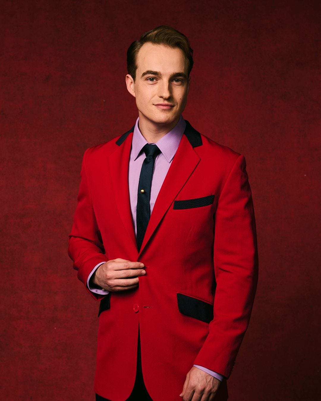 A man - Toby Miles - wearing a red blazer, purple shirt, and black tie against a red backdrop A man - Toby Miles - wearing a red blazer, purple shirt, and black tie against a red backdrop