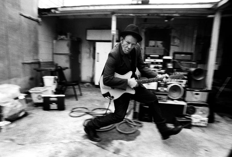 A black and white photo of a man in a suit and hat holding an electric guitar with a bank of speakers behind him. He is looking toward the camera and slightly down as he steps or possibly leaps forward. His shoes and the objects in the background of the photo are blurred, indicating that he is in motion. A black and white photo of a man in a suit and hat holding an electric guitar with a bank of speakers behind him. He is looking toward the camera and slightly down as he steps or possibly leaps forward. His shoes and the objects in the background of the photo are blurred, indicating that he is in motion.
