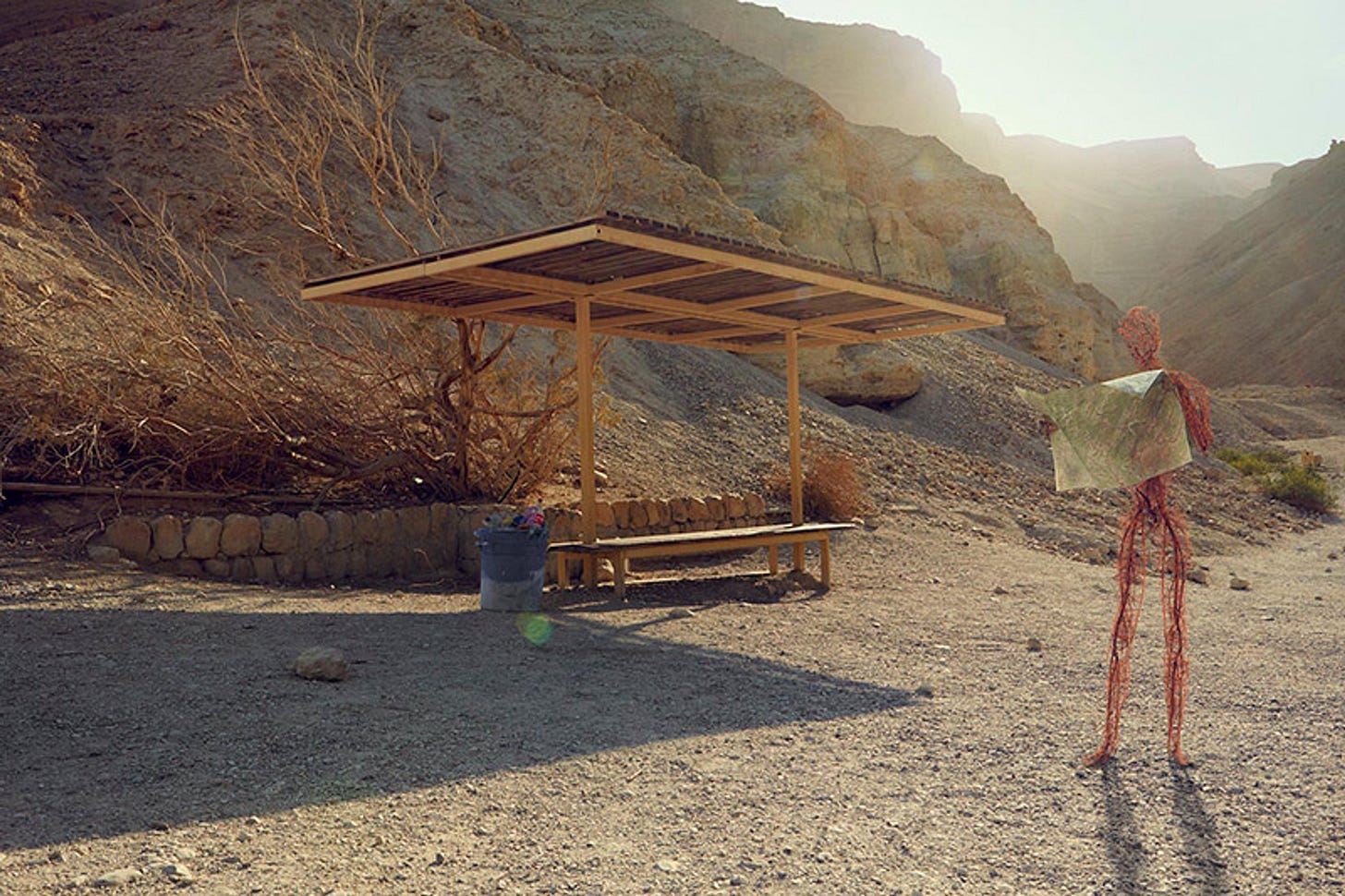 Photo of a wire sculpture figure holding a map in a desert landscape with a wooden shelter and hills in the background.