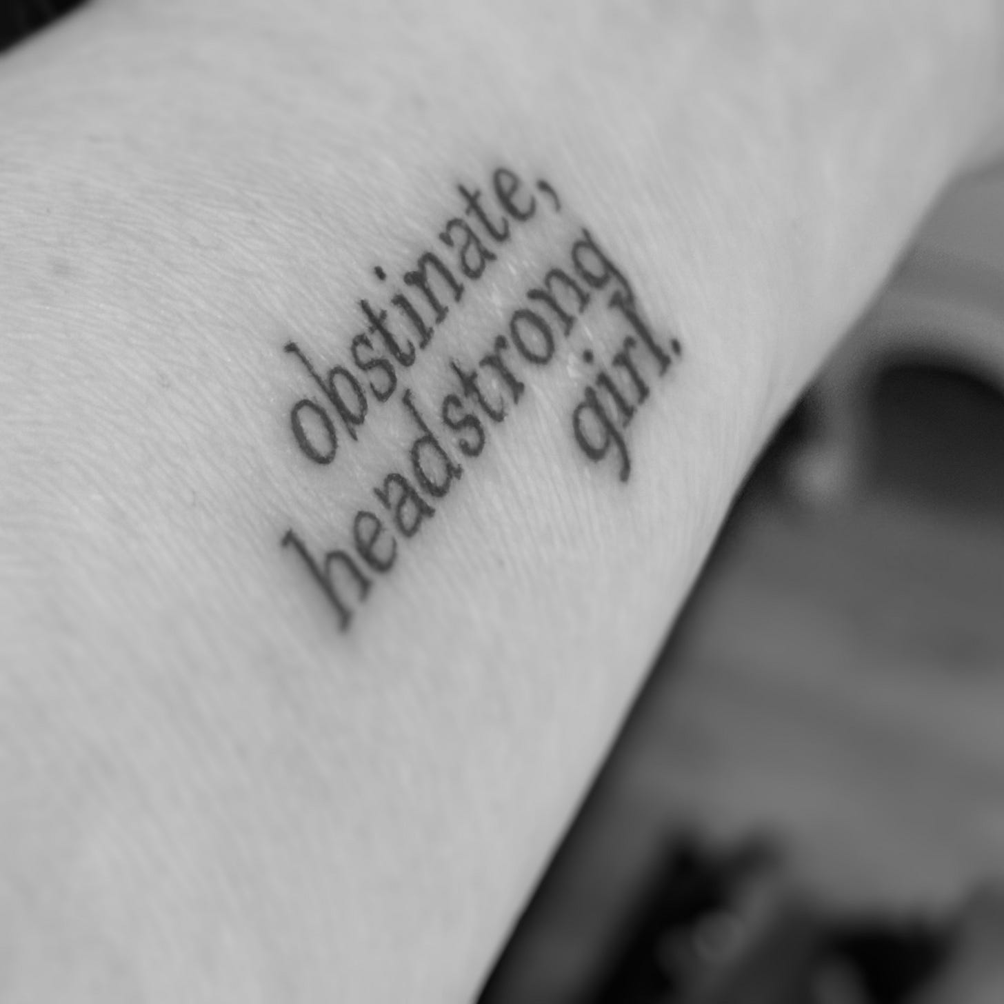 Black and white photograph of a tattoo on my arm reading 'obstinate headstrong girl' in a serif typeface.