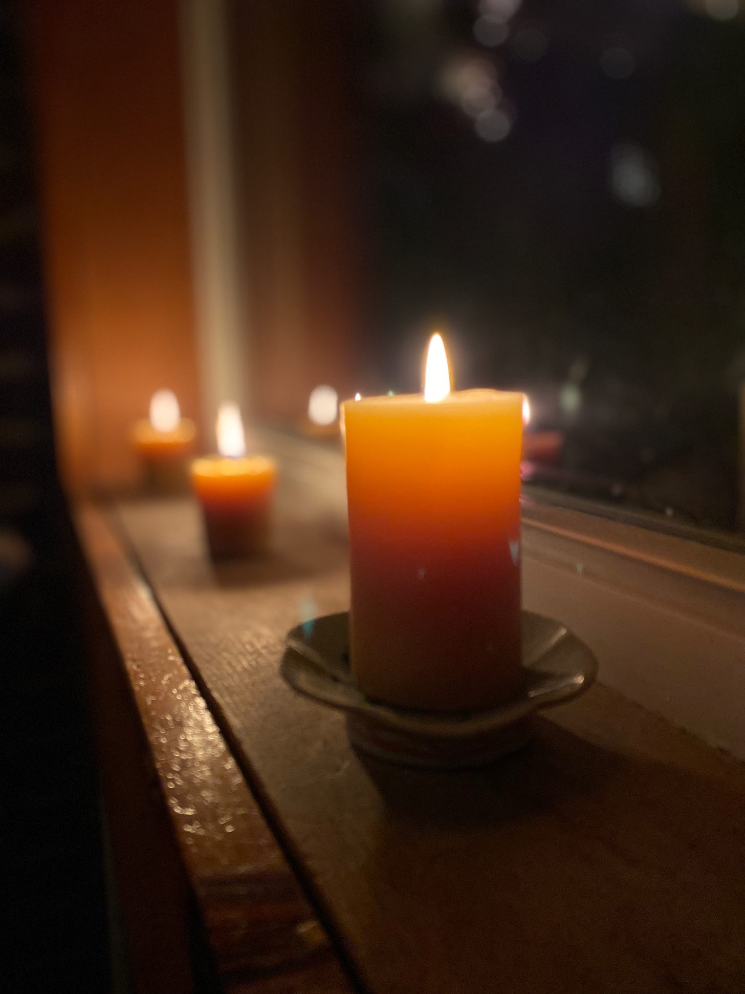 A lit beeswax candle on a windowsill. The window is dark, and several smaller candles, also lit, are blurred out in the background.
