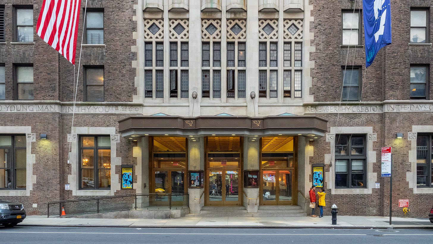 A photo of the exterior of the 92nd Street Y in New York City. There is an American flag and a flag with their logo hanging.