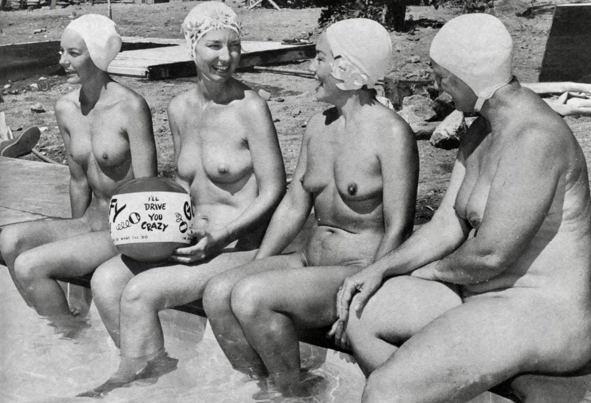 A black and white photograph of four nude women wearing swim caps sitting at the edge of a pool, laughing and holding a beach ball, at what appears to be a nudist resort.