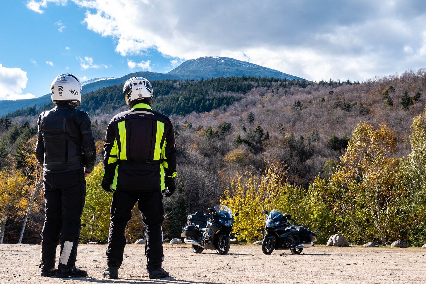 Tow motorcycle riders in full riding gear (including helmets) look up at a massive mountain, Mount Washington in New Hampshire. The mountain is bald up top and there appear to be patches of snow above the treeline. Further down the mountain are evergreens, and below, near the motorcycles, are trees with golden leaves.