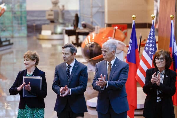 Four individuals in formal attire stand clapping. Flags and a large orange vehicle are visible in the background.