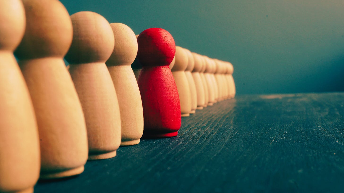 Red wooden figure standing out among a row of beige figures, symbolizing individuality and diversity in a leadership mosaic.