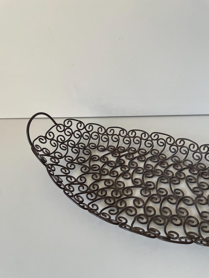 May include: A decorative metal leaf-shaped basket with a dark brown finish. The basket features a scrolled design throughout the body and a curved handle. The basket is designed to hold items and has a unique, artistic appearance.