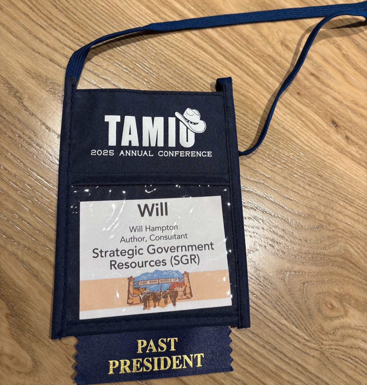 Close-up photo of a navy blue name badge holder from the 2025 TAMIO Annual Conference. The badge displays the name “Will Hampton,” with the title “Author, Consultant” and organization “Strategic Government Resources (SGR).” A graphic with cattle and the words “Saddle Up” appears at the bottom of the badge. A dark blue ribbon reading “PAST PRESIDENT” is attached to the holder. The badge is resting on a light wood surface.
