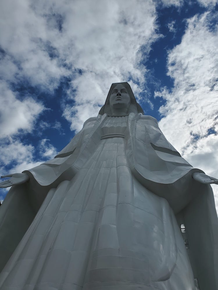 A white metal sculpture of the Virgin Mary looms overhead with her arms in an open gesture, a blue and clouded sky behind her.