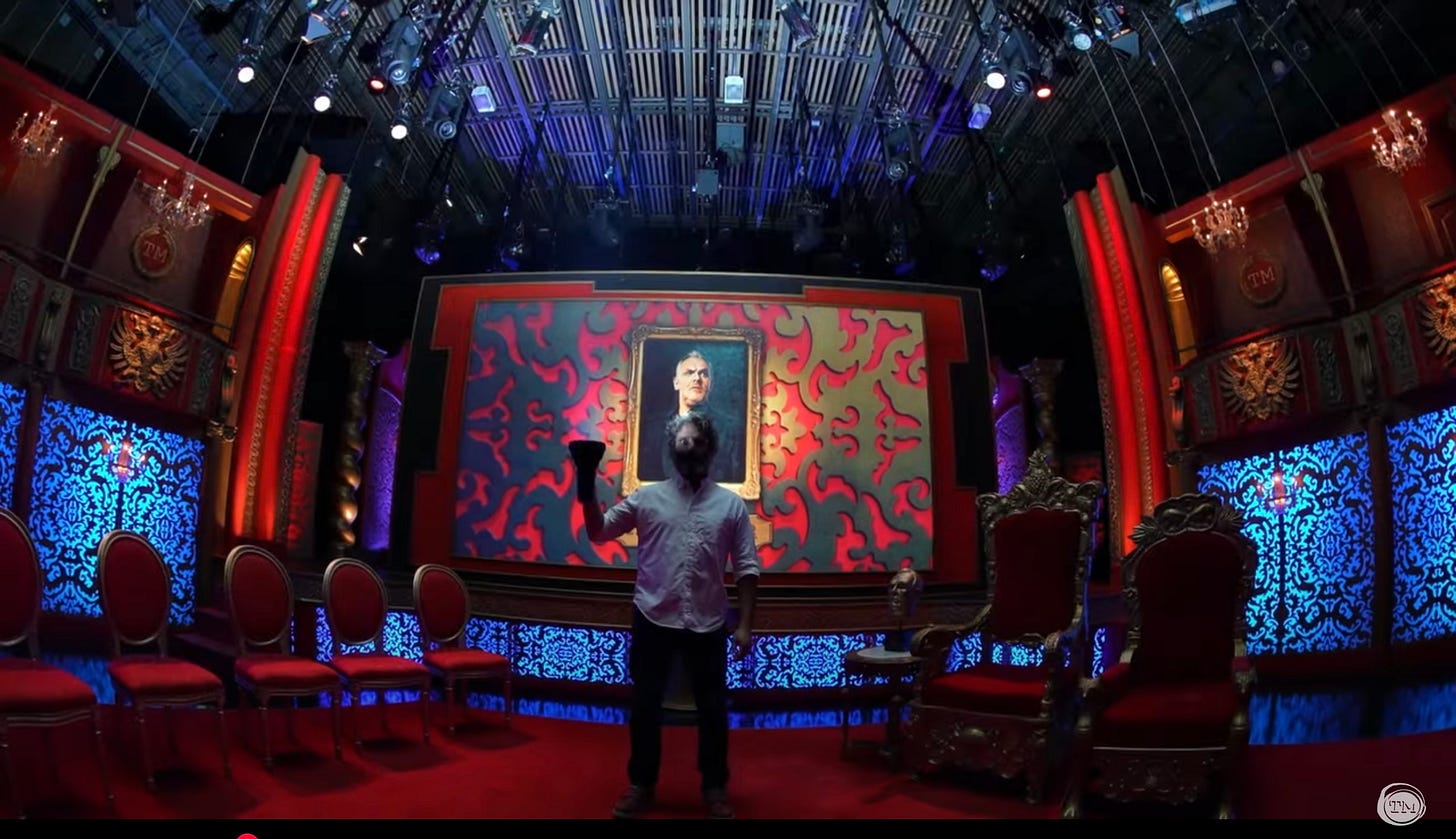 Jason Mantzoukas stands in the Taskmaster studio, holding a 200,000-lumen flashlight.