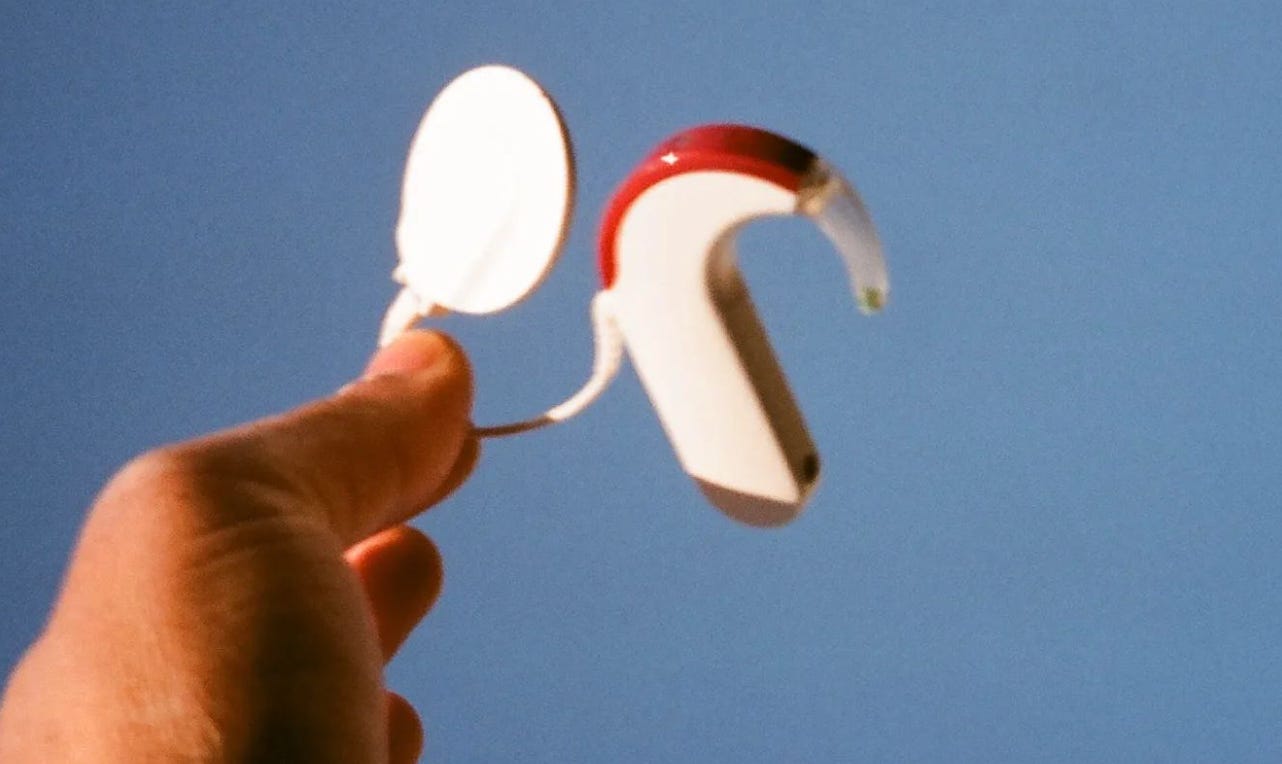 A hand holds the transmitter and processor, the external parts, of a Cochlear implant system against a blue sky in a grainy color photo.