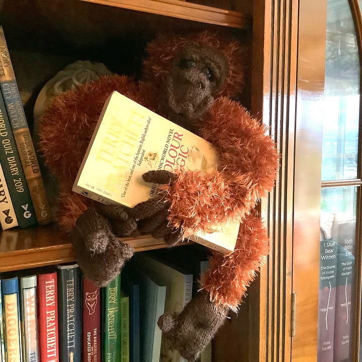 A stuffed toy orangutan in a bookcase with Terry Pratchett books and ornaments. 