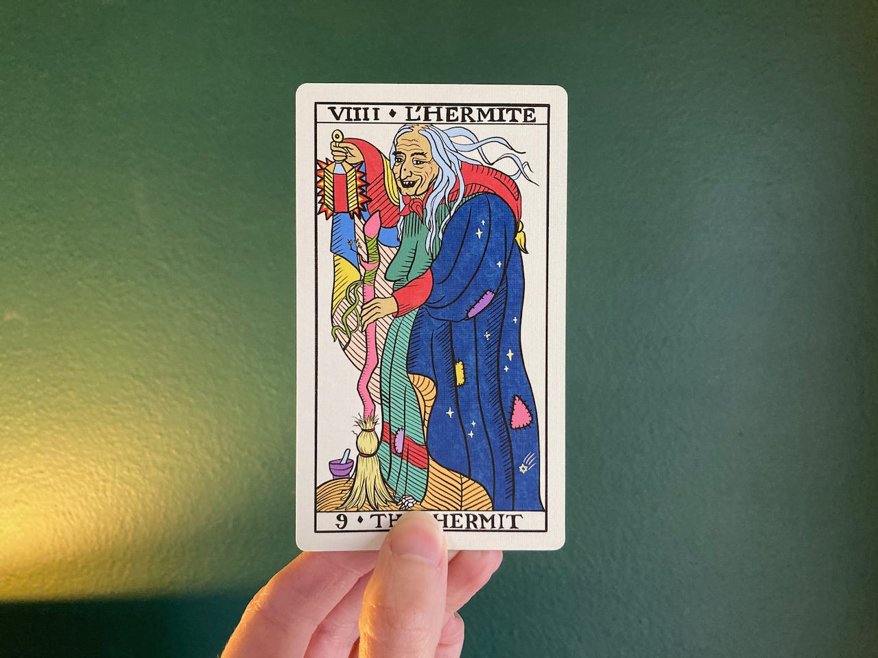 The Hermit from The Gay Marseille Tarot shows a woman with a wrinkled face and flyaway grey hair holding a shining lantern and a broom. Her patched cloak is decorated with stars, a mortar and pestle sit at her feet, and a skeletal foot of bones peeks from beneath her skirts.