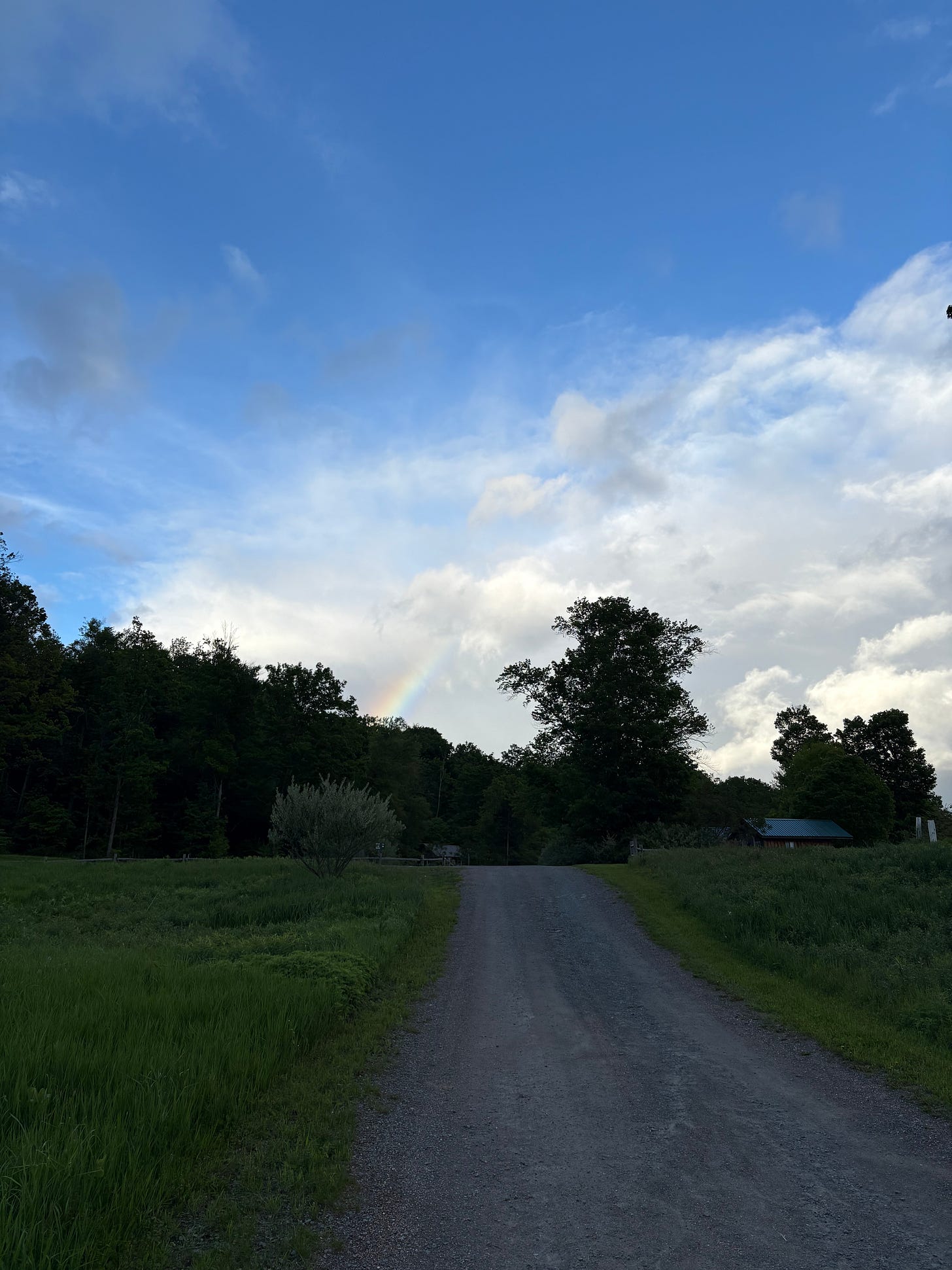 A bit of a rainbow is visible in a cloudy blue sky. Below is a gravel path surrounded by green grass and woods.