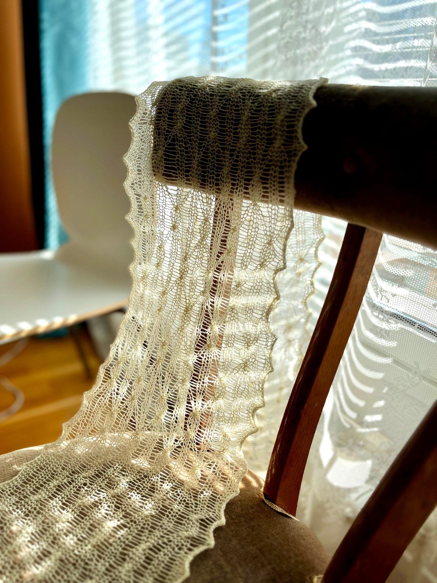 A finished cream lace scarf hanging vertically against a sage green wall. The full length is visible, showing the repeating elongated diamond pattern, scalloped side edges, and the fine drape of the single-ply wool.