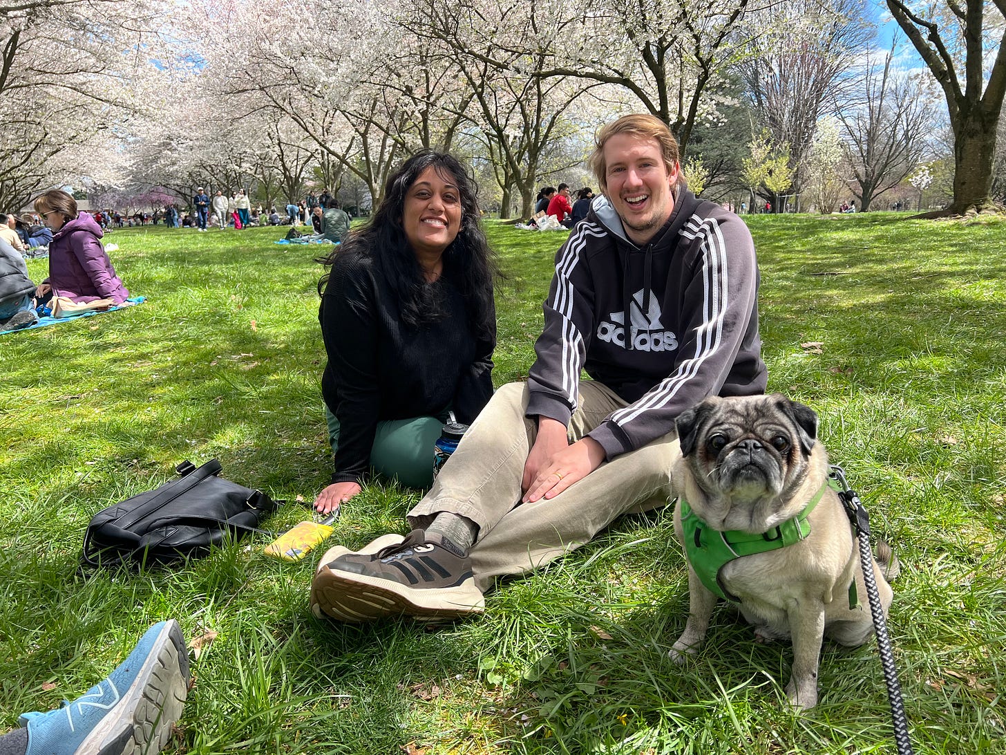 Photo of smiling Indian woman (me) and her husband white husband sitting in the grass under cherry trees with a pug. Photo of smiling Indian woman (me) and her husband white husband sitting in the grass under cherry trees with a pug.