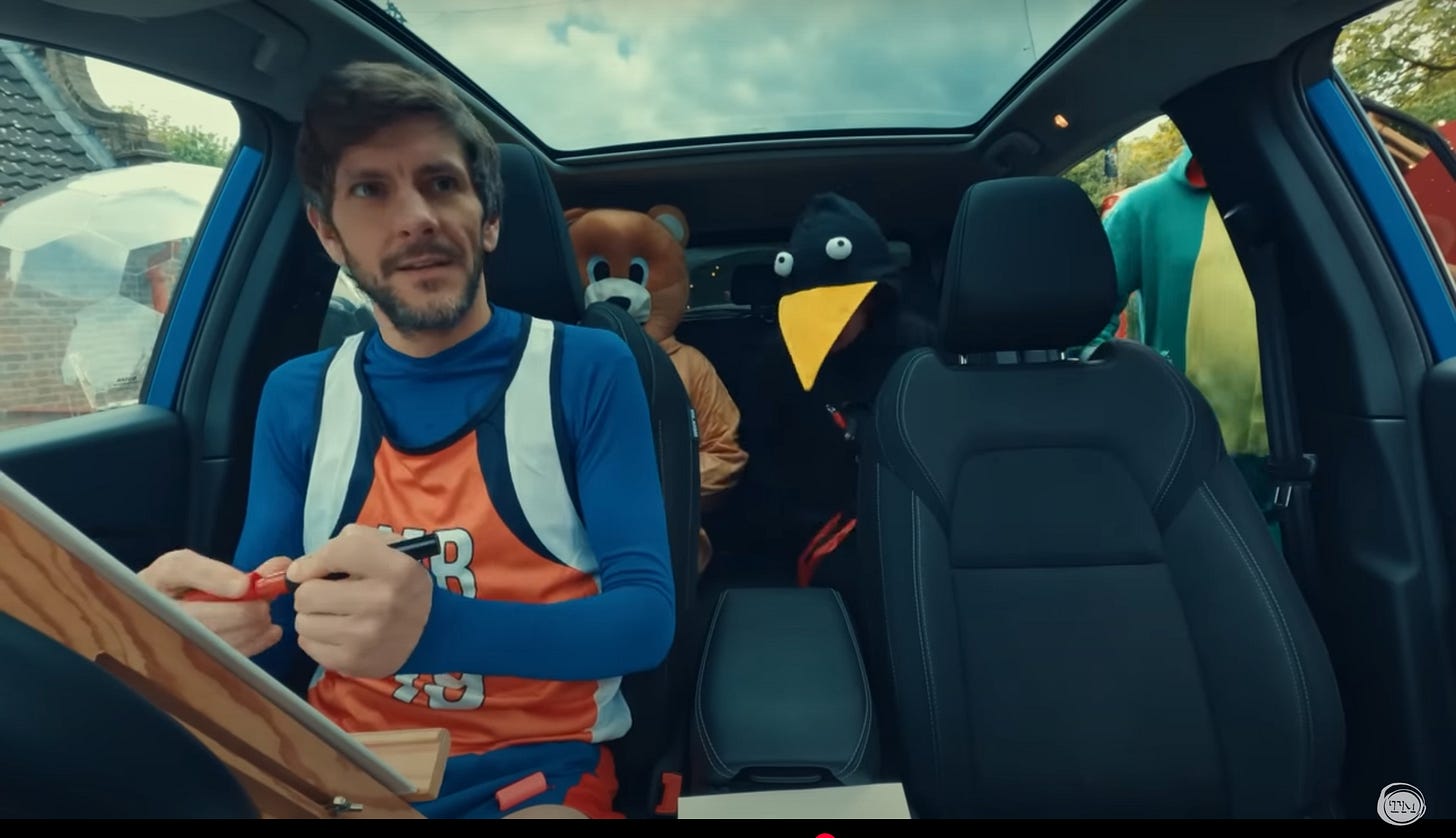 Mathew Baynton sits in a car with an easel in front of him. In the backseat are two people, one dressed as a giant teddy bear and one dressed as a giant crow.
