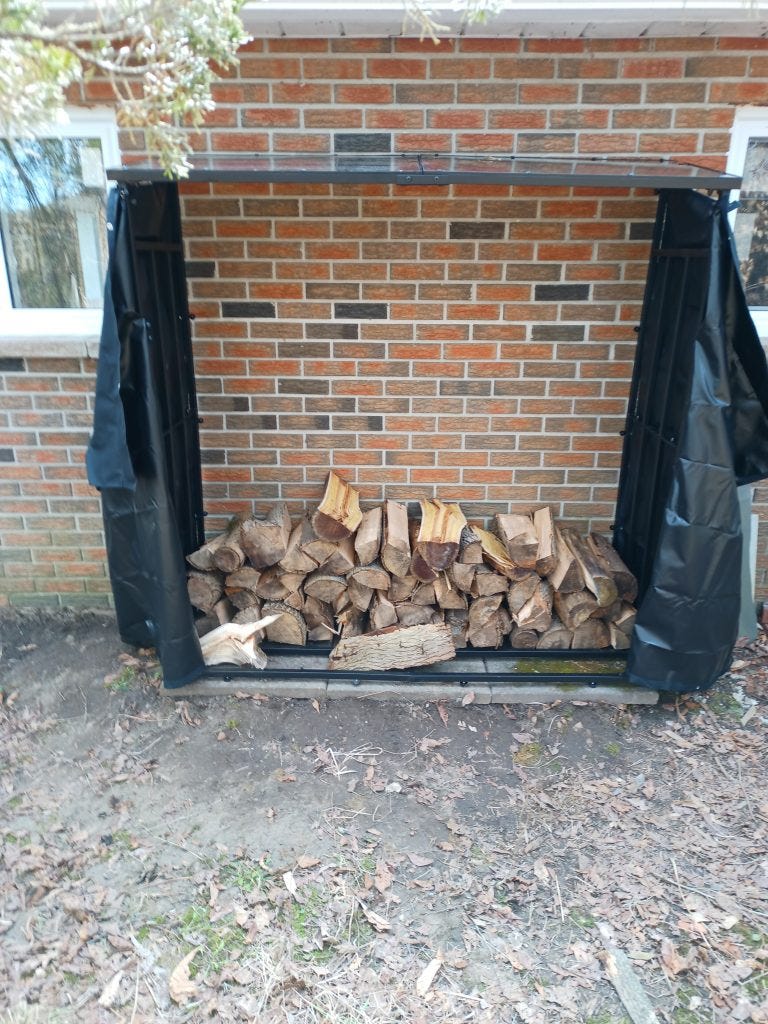A large wood storage rack about 2m wide and high, and about 600mm deep, set on concrete tiles and against a brick wall. Black covers hang open at each end.