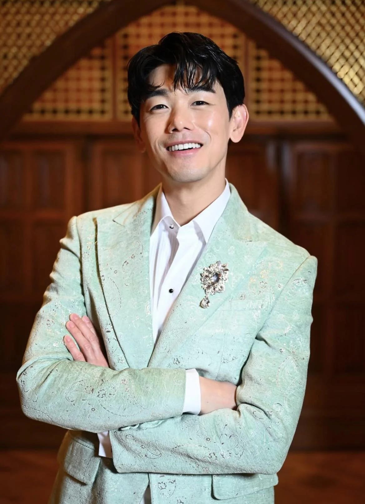 Eric in a mint green brocade suit jacket with silver brooch, white collared shirt with black buttons, arms crossed, smiling.