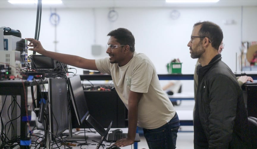 Two men in foreground review a device showing waveforms; a man in the background is mostly hidden. Two men in foreground review a device showing waveforms; a man in the background is mostly hidden.