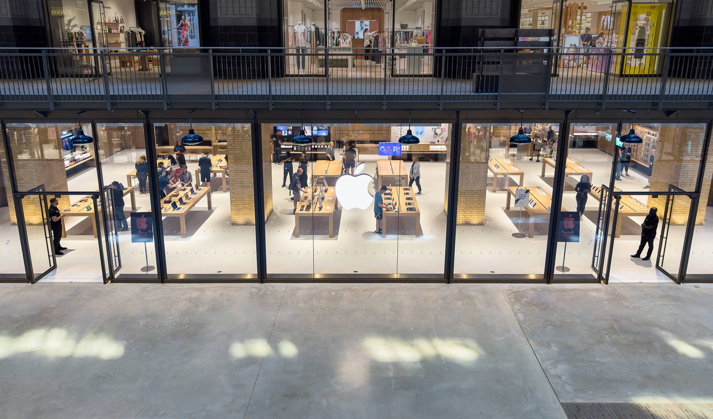 The storefront of Apple Battersea from an upper balcony. Customers move about the tables inside. The storefront of Apple Battersea from an upper balcony. Customers move about the tables inside.