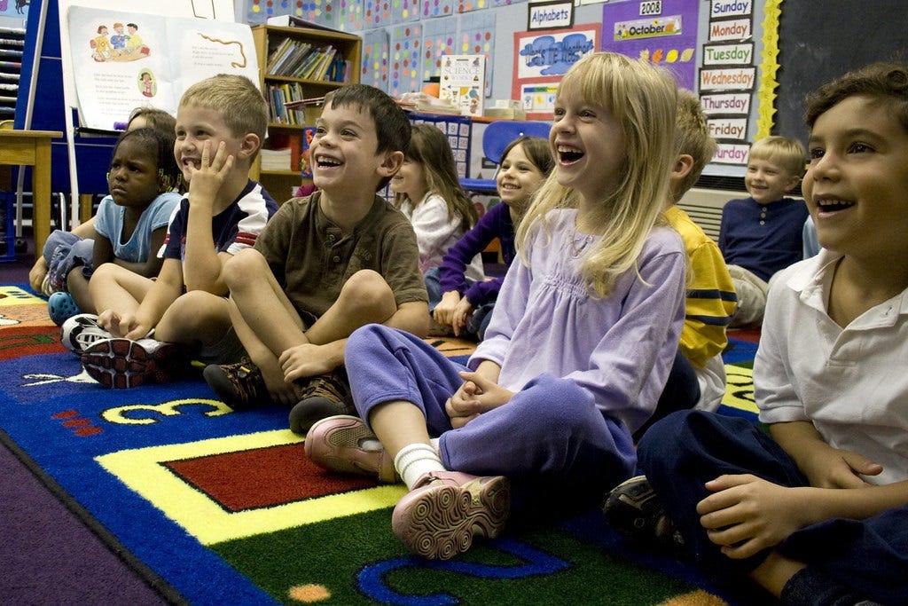 Group of kindergarteners sitting on bright, colorful carpet in a classroom, laughing at something we can't see. One child looks perturbed for unknown reasons. Group of kindergarteners sitting on bright, colorful carpet in a classroom, laughing at something we can't see. One child looks perturbed for unknown reasons.