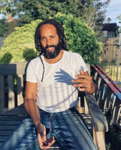 Photo of The Roots farmer, a Black man with a beard and dreadlocks. He is sat on a bench wearing a white t shirt and blue jeans. Photo of The Roots farmer, a Black man with a beard and dreadlocks. He is sat on a bench wearing a white t shirt and blue jeans.