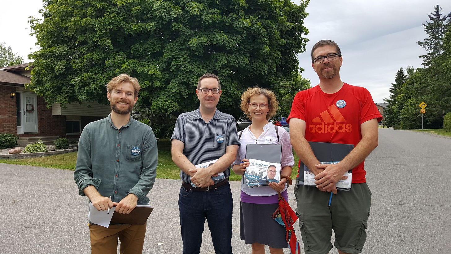Four people standing on a street with a house and tree in the backround. Each wearing a blue button, some holding flyers or clipboards