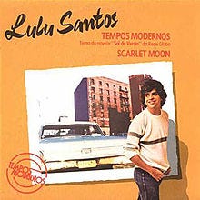 foto da capa do disco tempos modernos de lulu santos. fundo laranja, dizeres em preto e vermelho. o artista encontra-se em primeiro plano, com calça jeans e blusa de manga comprida. ao fundo, um carro e dois prédios.