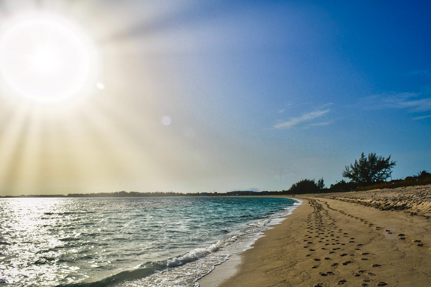 The bright sun shining on the beach with turquoise water and footprints in the sand along the shore