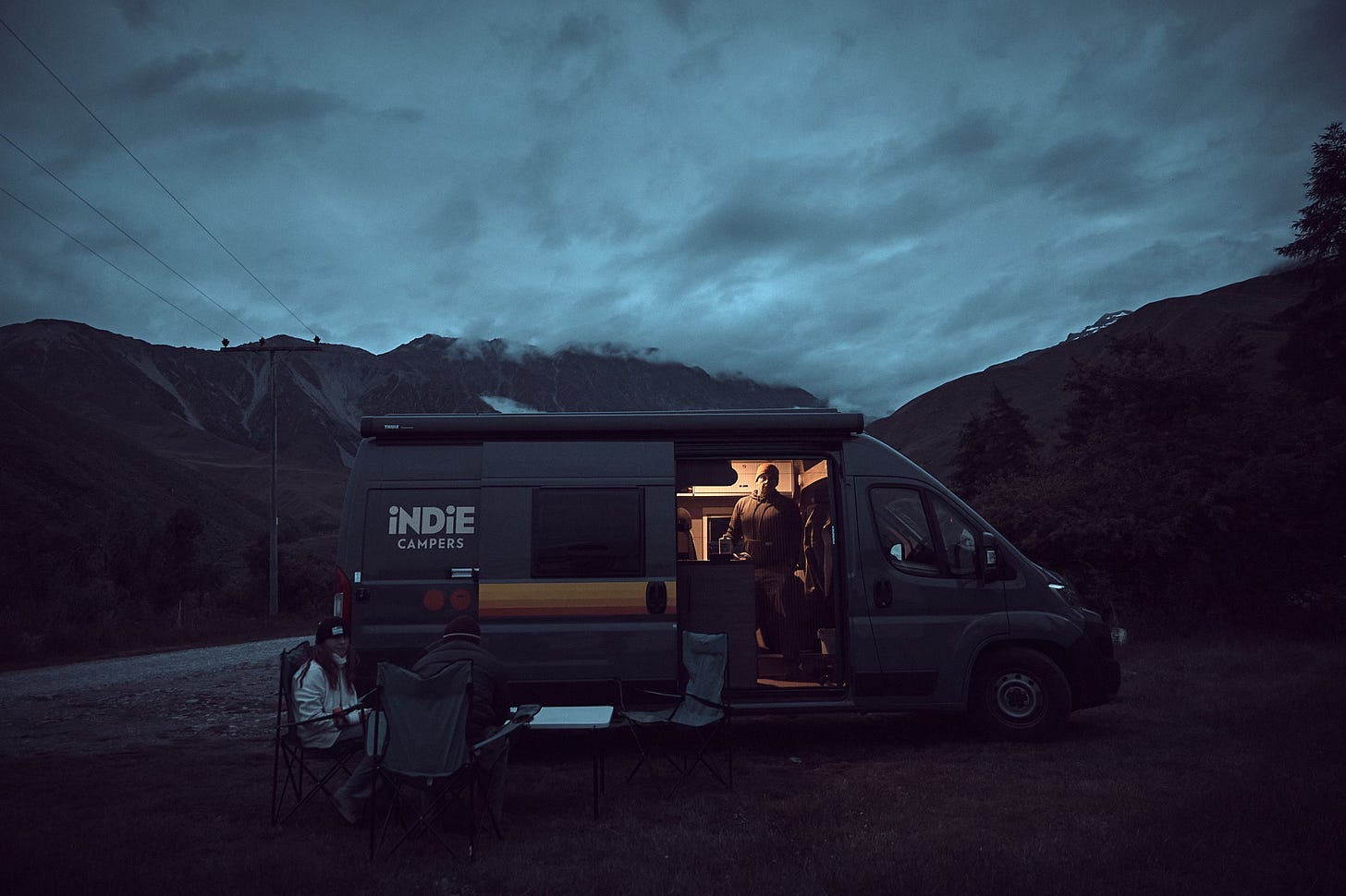 Jon standing, lit inside camper van as Maddie and Dylan sit, rugged up outside, the mountains around Mt Cook in the background