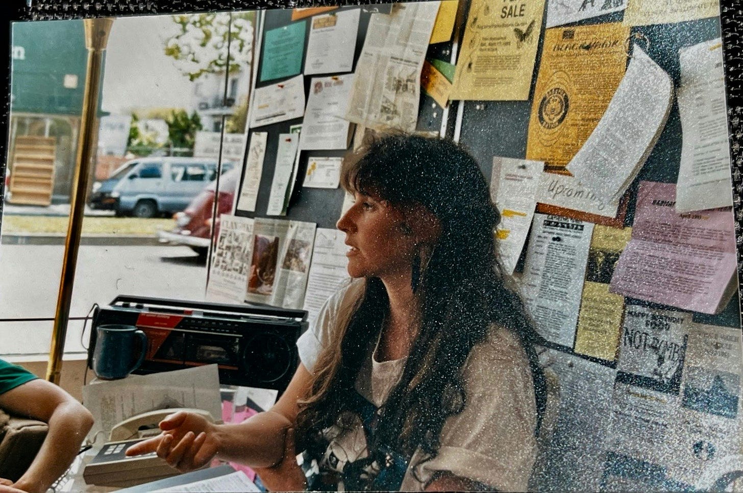 a young woman with lots of dark hair, wearing a loose-fitting white tshirt, sitting in front of a bulletin board covered with flyers.