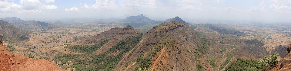A wide panorama from a hill viewpoint at Matheran, looking out across ridgelines and distant lowlands, with a railway line visible along the ridge.