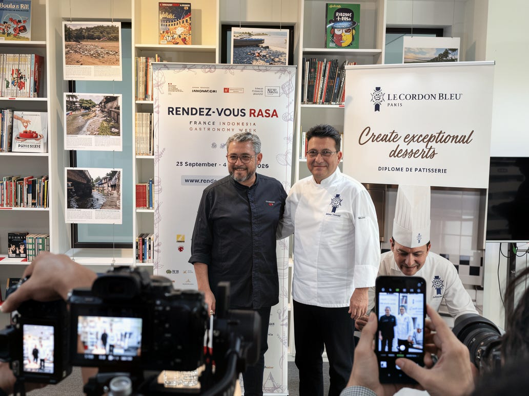 Two chefs pose side by side in front of a Rendez-vous Rasa France Indonesia Gastronomy Week banner, one in a dark jacket and one in a white Le Cordon Bleu Paris jacket, while photographers shoot them Two chefs pose side by side in front of a Rendez-vous Rasa France Indonesia Gastronomy Week banner, one in a dark jacket and one in a white Le Cordon Bleu Paris jacket, while photographers shoot them