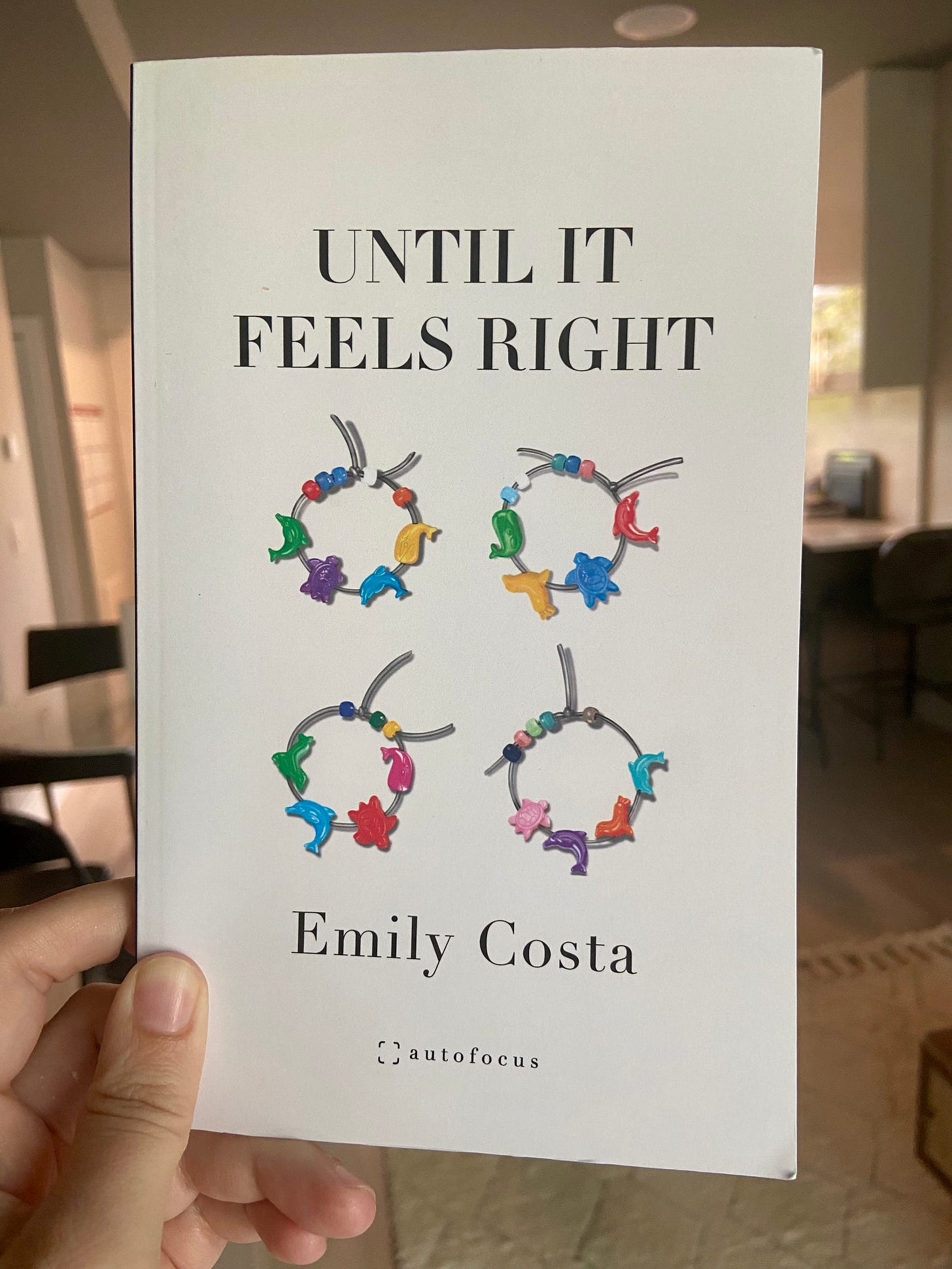 White hand holding a white book called "Until It Feels Right" by Emily Costa