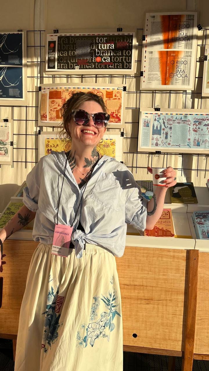 Mulher branca de pé na frente de uma bancada de livros, usando saia branca, cabelo preso, camisa azul com as mangas enroladas, sorrindo e usando óculos escuros